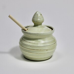 May include: A light green ceramic honey pot with a ribbed body and a matching lid featuring a pointed finial. A light-colored wooden honey dipper is inserted through a small notch in the lid, resting against the side of the pot.