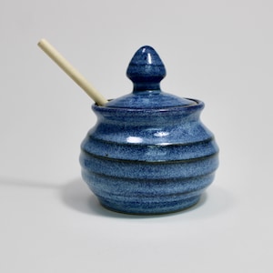 以下が含まれることがあります： 丸みを帯びた青い陶器製のハニーポット。本体には溝があり、先端が尖ったつまみ付きの蓋が被せられています。明るい色の木製ハニーディッパーがポットに差し込まれ、蓋の開口部に斜めに立てかけられています。