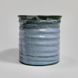 May include: A handmade ceramic tumbler with a textured, ridged surface. The stoneware vessel features a gradient of blue and green hues, with a darker rim and lighter body. The pottery is suitable for beverages.