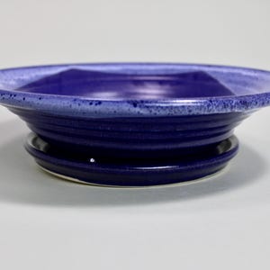 Może przedstawiać: Ręcznie robiona ceramiczna miska z głębokim indygowym szkliwem. Miska ma szeroki, lekko rozszerzany brzeg i żebrowaną podstawę. Wnętrze jest w jaśniejszym odcieniu niebieskiego, tworząc kontrast. Miska ma średnicę około 20 cm.