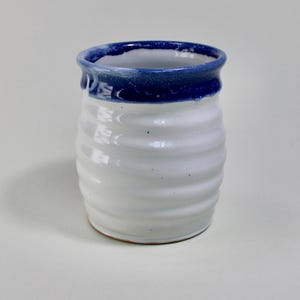 May include: A handcrafted ceramic tumbler. The white body has horizontal ridged detailing, and the rim is dark blue. The interior is white, and the base is unglazed. This mug is suitable for beverages. The height is approximately 10 cm.