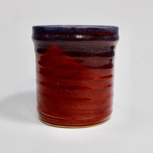 Pode incluir: Uma caneca cilíndrica de cerâmica com um esmalte brilhante que transita de um roxo profundo e escuro na borda para um vermelho vibrante e saturado no corpo. A superfície apresenta sulcos horizontais subtis e um acabamento liso e reflexivo.