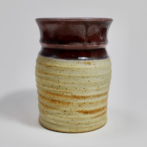 Puede incluir: Un jarrón de cerámica hecho a mano con forma cilíndrica. La sección superior es de color burdeos oscuro, mientras que el cuerpo es de color beige moteado con ranuras horizontales y detalles marrones. El jarrón tiene un borde ligeramente ensanchado.