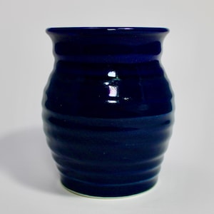 May include: A deep navy blue ceramic vase with a glossy finish and a series of horizontal ridges encircling the body. The vase features a wide, flared rim and a rounded, bulbous shape that tapers toward a flat base, reflecting light on its surface.