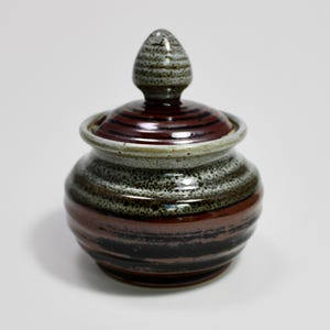 May include: A ceramic jar with a lid, featuring a striped design in brown, black, and grey. The lid has a rounded top and a matching colour scheme. The jar is a decorative item, suitable for storage or display.