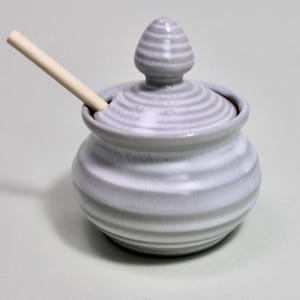 May include: A gray ceramic sugar bowl with a lid and a small wooden spoon. The bowl has a ribbed design and a matching lid with a knob. The spoon rests inside the bowl. The sugar bowl is on a light green surface.