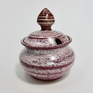 May include: A ceramic sugar bowl with a lid and a small opening for a spoon. The bowl is a speckled white and burgundy colour with burgundy stripes. The lid has a decorative finial.
