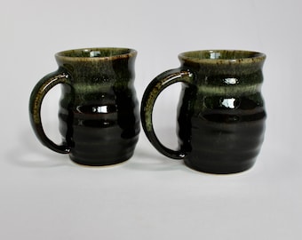 May include: Two handmade ceramic mugs with a dark green and black glaze. Each mug has a large handle and a ridged body. The mugs are suitable for coffee, tea, or other beverages. The mugs are displayed on a white background.