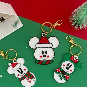 May include: Three acrylic keychains featuring white mouse-shaped characters with holiday-themed accessories. One wears a red Santa hat and striped scarf, another a red hat, and the third a green cape with red polka dots, all with gold clasps.