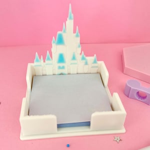 Puede incluir: Un sujetapapeles blanco en forma de castillo con detalles azules, que contiene un bloc de notas gris. Tambi&eacute;n hay un bol&iacute;grafo con la palabra "Kaleedos" y un dise&ntilde;o de Mickey Mouse. El fondo es rosa.