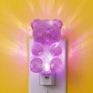 May include: A purple, translucent, gummy bear-shaped night light plugged into a white wall outlet. The bear has a textured, crystalline surface and emits a bright, glowing purple light that radiates outward against the yellow wall.