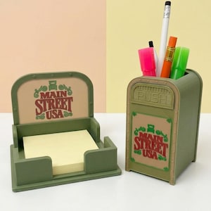 May include: A sage green desk organizer set featuring a square sticky note holder and a matching pencil cup shaped like a trash bin. Both items display the text Main Street USA in red lettering with green decorative flourishes.