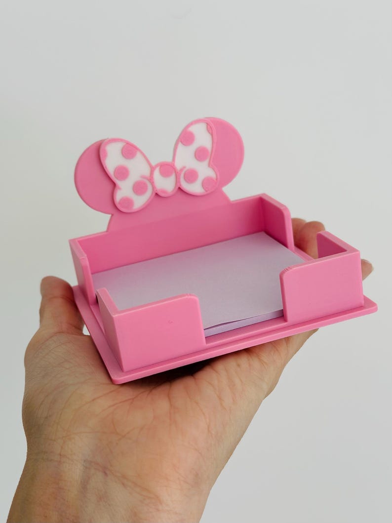 Puede incluir: Un organizador de escritorio rosa con forma de lazo con lunares blancos y dos orejas redondeadas. El organizador contiene una pila de papel de notas de color p&uacute;rpura claro. El art&iacute;culo se sostiene en la mano de una persona.