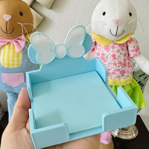 Puede incluir: Un soporte para notas adhesivas azul claro con un adorno en forma de lazo. El soporte es de madera y tiene forma rectangular. El fondo presenta dos peluches de conejos de Pascua.