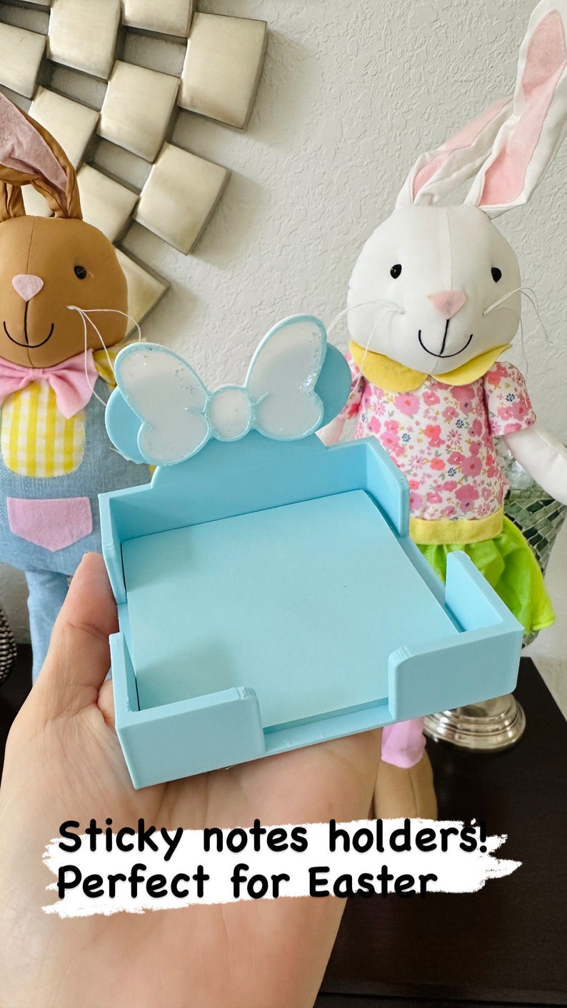 Puede incluir: Un soporte para notas adhesivas azul claro con un adorno en forma de lazo. El soporte es de madera y tiene forma rectangular. El fondo presenta dos peluches de conejos de Pascua.