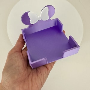 Puede incluir: Un organizador de escritorio morado con forma de rat&oacute;n con un lazo blanco. El organizador se sostiene en una mano y tiene una base rectangular con lados elevados. Las orejas y el lazo del rat&oacute;n son blancos.