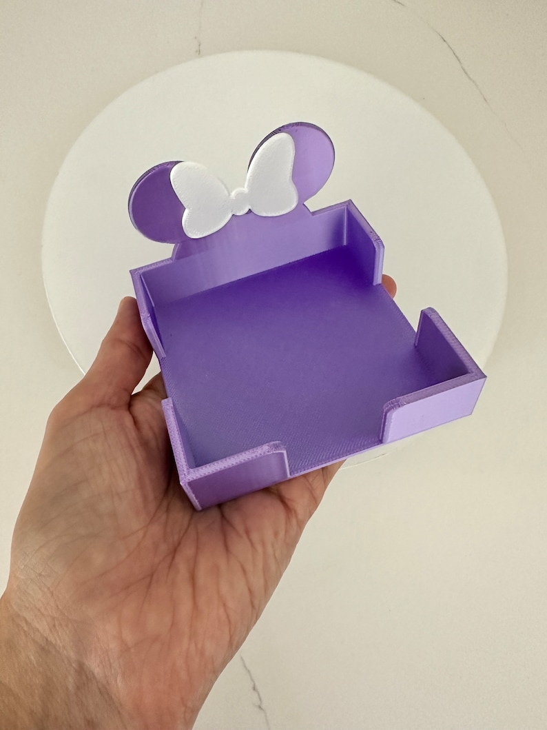Puede incluir: Un organizador de escritorio morado con forma de rat&oacute;n con un lazo blanco. El organizador se sostiene en una mano y tiene una base rectangular con lados elevados. Las orejas y el lazo del rat&oacute;n son blancos.