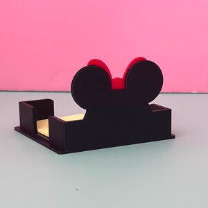 Puede incluir: Un organizador de escritorio negro con forma de rat&oacute;n y un lazo rojo. El organizador tiene una base rectangular con secciones verticales para sujetar notas adhesivas. El fondo es de color rosa liso.