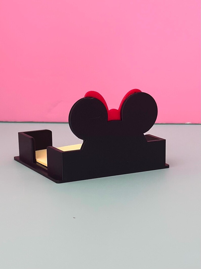 Puede incluir: Un organizador de escritorio negro con forma de rat&oacute;n y un lazo rojo. El organizador tiene una base rectangular con secciones verticales para sujetar notas adhesivas. El fondo es de color rosa liso.
