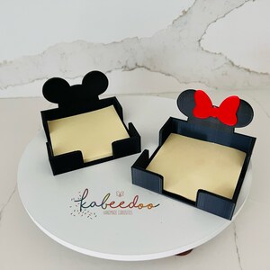 Puede incluir: Dos sujetapapeles negros, uno con un dise&ntilde;o de oreja de Mickey Mouse y el otro con un dise&ntilde;o de oreja de Minnie Mouse y un lazo rojo. Cada soporte contiene una pila de notas adhesivas amarillas. Los soportes est&aacute;n sobre una superficie blanca.
