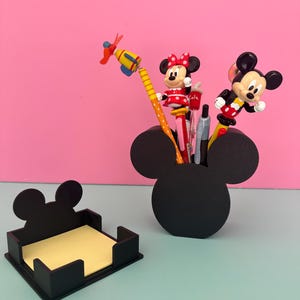Puede incluir: Organizador de escritorio negro con forma de Mickey Mouse y un portanotas a juego. El organizador contiene bol&iacute;grafos y l&aacute;pices con adornos de los personajes de Mickey y Minnie Mouse. El portanotas contiene notas adhesivas amarillas. El fondo es rosa y azul claro.