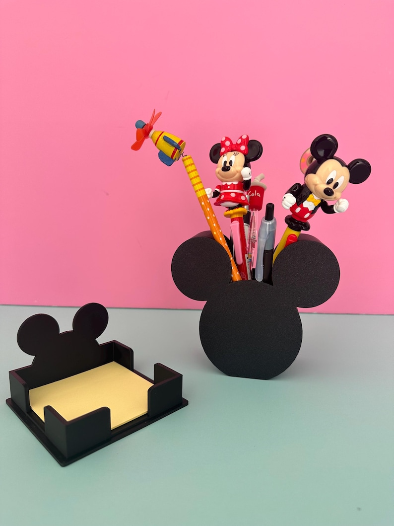 Puede incluir: Organizador de escritorio negro con forma de Mickey Mouse y un portanotas a juego. El organizador contiene bol&iacute;grafos y l&aacute;pices con adornos de los personajes de Mickey y Minnie Mouse. El portanotas contiene notas adhesivas amarillas. El fondo es rosa y azul claro.