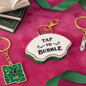 May include: Keychains with gold-colored hardware and charms. One keychain features a map of Oz with a witch's hat, another has a fan-shaped charm with the text "TAP TO BUBBLE", and a third has a small shoe charm. Green ribbon and miniature books are also present.