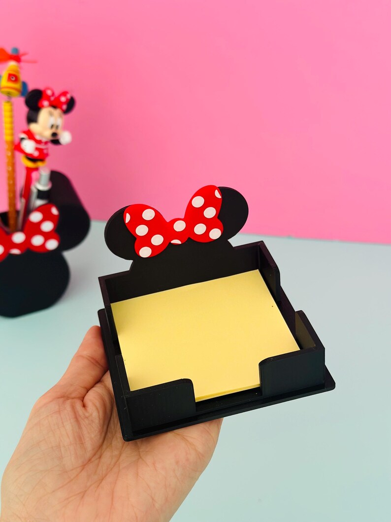 Puede incluir: Organizador de escritorio cuadrado negro con un dise&ntilde;o de lazo de lunares rojos y blancos. El organizador contiene una pila de notas adhesivas amarillas. Un portal&aacute;pices con el tema de Minnie Mouse es visible en el fondo.