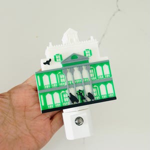 May include: A decorative night light featuring a green and white haunted house design. The house has a detailed facade with windows and a gray roof. Black silhouettes of figures are visible. The night light plugs into a standard wall outlet.