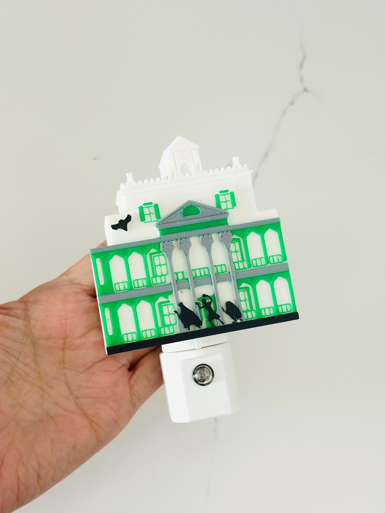 May include: A decorative night light featuring a green and white haunted house design. The house has a detailed facade with windows and a gray roof. Black silhouettes of figures are visible. The night light plugs into a standard wall outlet.