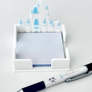 Puede incluir: Un portanotas blanco en forma de castillo con detalles azules, que contiene papel de notas azul claro. Un bol&iacute;grafo con la palabra "kabeedoo" y un dibujo de Mickey Mouse est&aacute; al lado del portanotas.