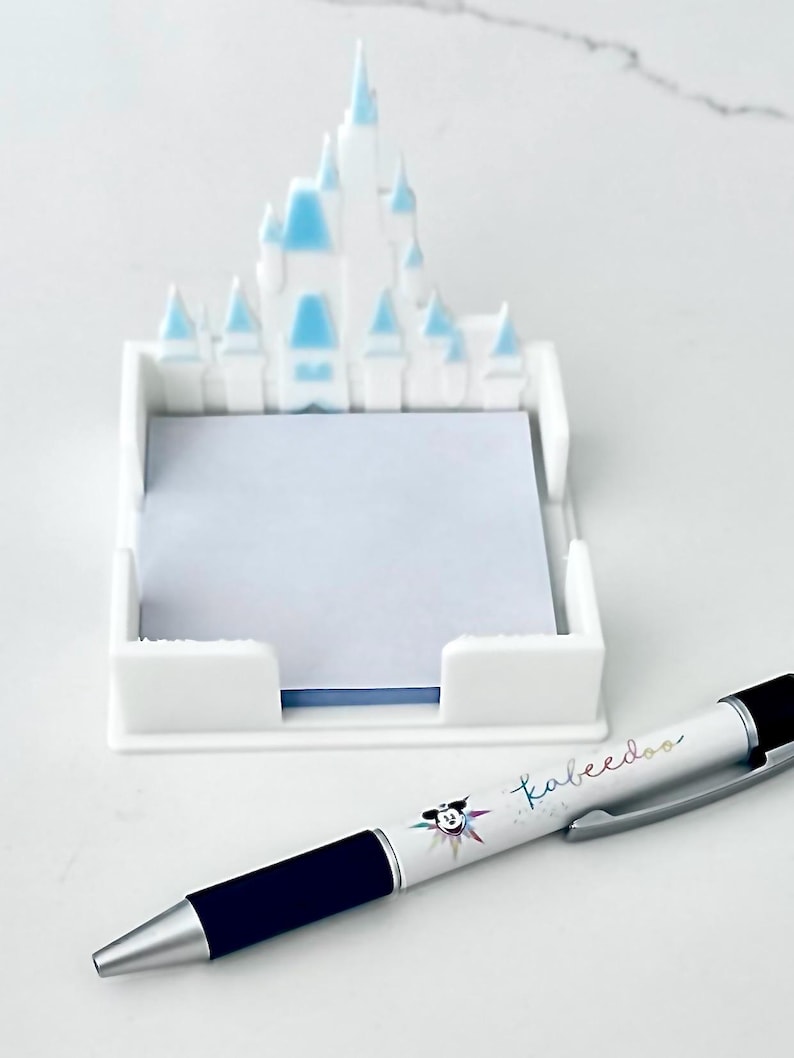 Puede incluir: Un portanotas blanco en forma de castillo con detalles azules, que contiene papel de notas azul claro. Un bol&iacute;grafo con la palabra "kabeedoo" y un dibujo de Mickey Mouse est&aacute; al lado del portanotas.