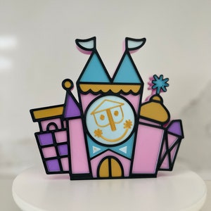 May include: A pink, blue, and yellow 3D printed castle with a smiling face on the clock tower. The castle is a stylized version of a famous theme park attraction.