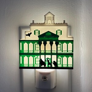 May include: A decorative night light shaped like a mansion, featuring a green and white color scheme. The mansion has multiple windows and a silhouette of figures in front. The night light plugs into a standard wall outlet.