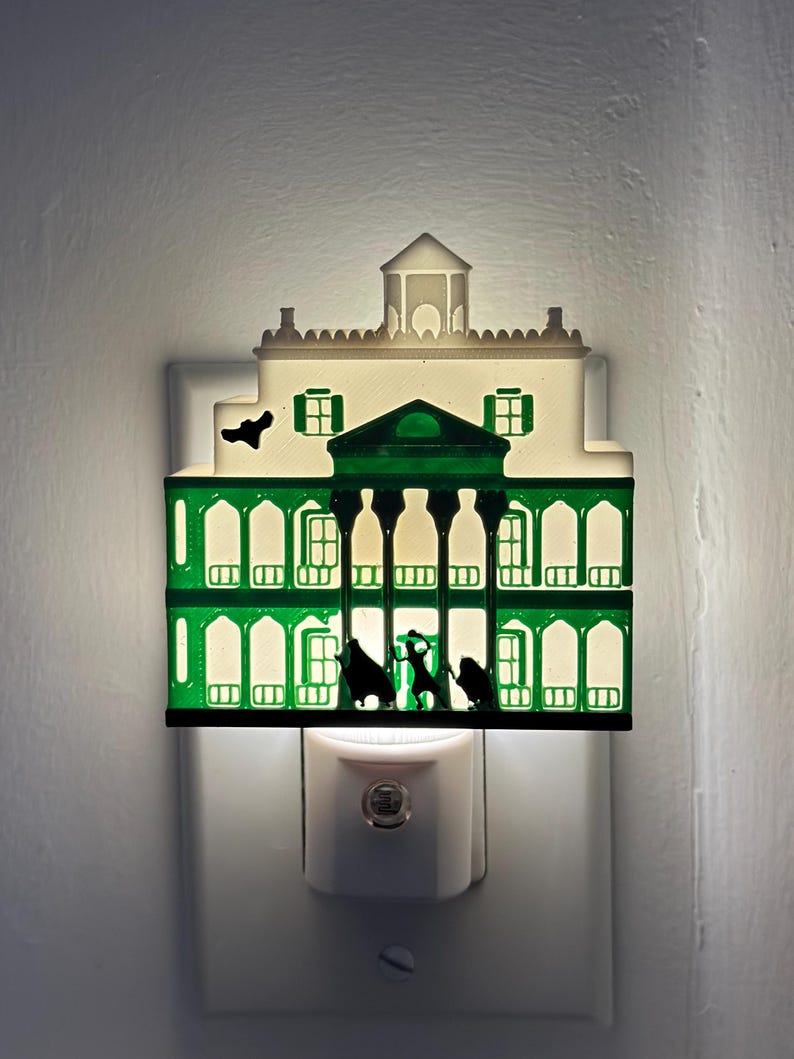 May include: A decorative night light shaped like a mansion, featuring a green and white color scheme. The mansion has multiple windows and a silhouette of figures in front. The night light plugs into a standard wall outlet.