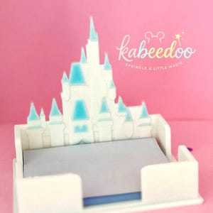 Puede incluir: Un organizador de escritorio blanco en forma de castillo con detalles en azul claro en las torres y un interior gris. El organizador tiene el texto "kabeedoo Sprinkle a little magic" en una fuente caprichosa sobre un fondo rosa.