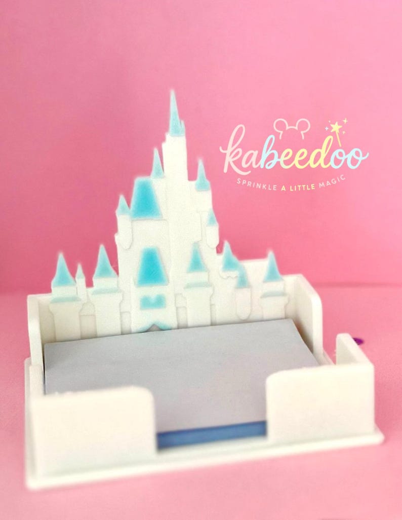 Puede incluir: Un organizador de escritorio blanco en forma de castillo con detalles en azul claro en las torres y un interior gris. El organizador tiene el texto "kabeedoo Sprinkle a little magic" en una fuente caprichosa sobre un fondo rosa.