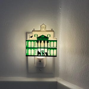 May include: A decorative night light shaped like a mansion, with a green and white color scheme. The night light is plugged into a wall outlet and emits a soft, warm glow. The mansion design features silhouettes of figures and architectural details.