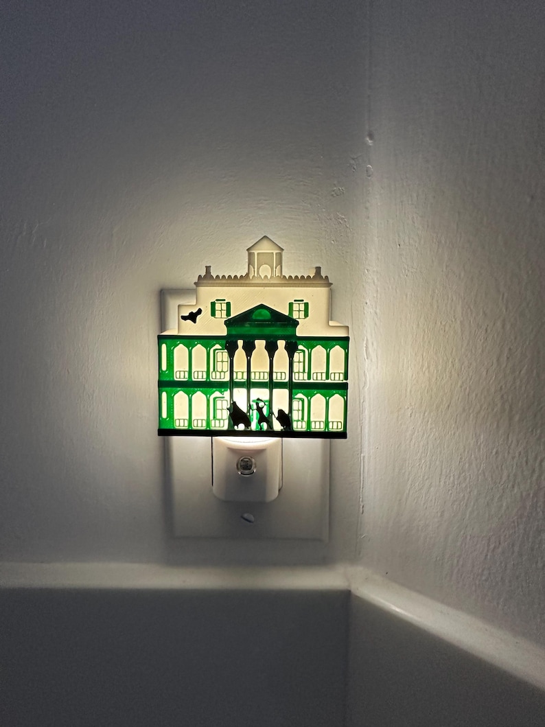 May include: A decorative night light shaped like a mansion, with a green and white color scheme. The night light is plugged into a wall outlet and emits a soft, warm glow. The mansion design features silhouettes of figures and architectural details.