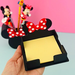 Puede incluir: Un organizador de escritorio negro con forma de cabeza de Minnie Mouse, con un lazo rojo de lunares blancos. El organizador contiene notas adhesivas amarillas. En el fondo hay figuras de Mickey y Minnie Mouse y l&aacute;pices.