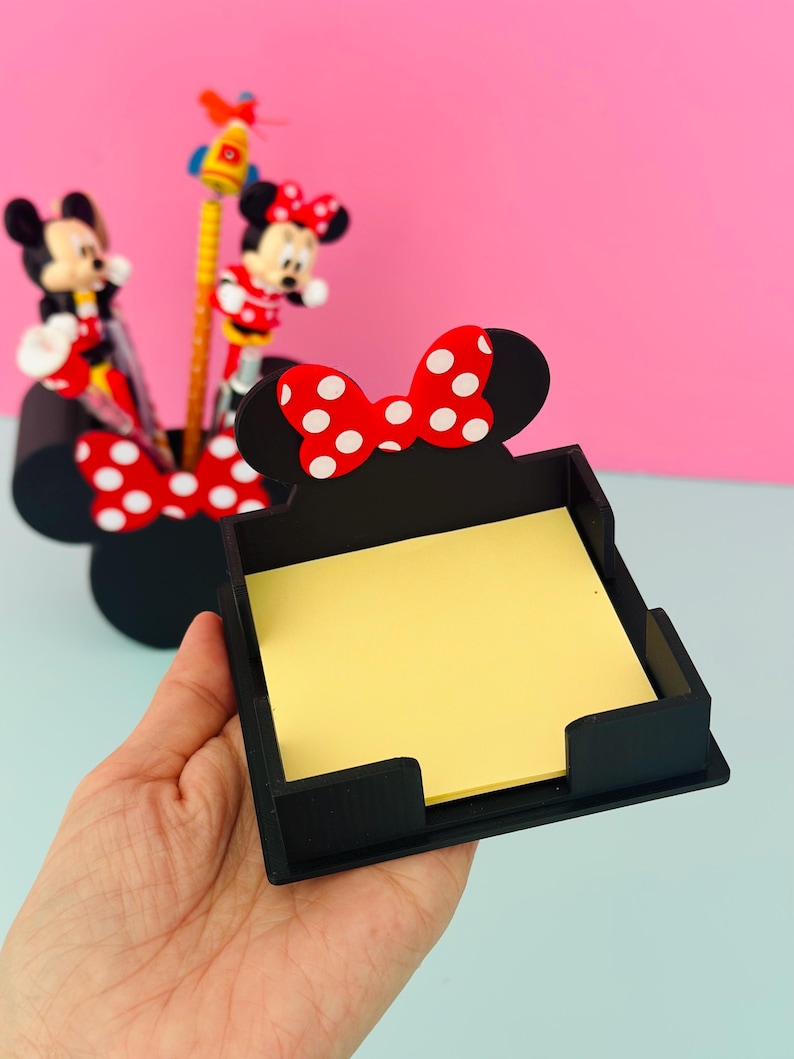 Puede incluir: Un organizador de escritorio negro con forma de cabeza de Minnie Mouse, con un lazo rojo de lunares blancos. El organizador contiene notas adhesivas amarillas. En el fondo hay figuras de Mickey y Minnie Mouse y l&aacute;pices.
