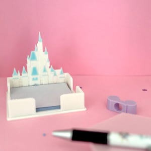 Puede incluir: Un organizador de escritorio blanco en forma de castillo con detalles en azul claro, que contiene una pila de papel de notas gris. Un bol&iacute;grafo negro y plateado y un peque&ntilde;o objeto morado tambi&eacute;n est&aacute;n sobre la superficie rosa.