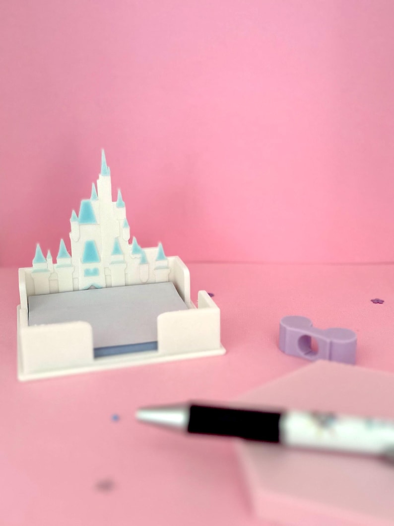 Puede incluir: Un organizador de escritorio blanco en forma de castillo con detalles en azul claro, que contiene una pila de papel de notas gris. Un bol&iacute;grafo negro y plateado y un peque&ntilde;o objeto morado tambi&eacute;n est&aacute;n sobre la superficie rosa.