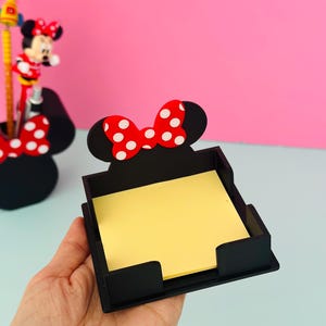 Puede incluir: Organizador de escritorio cuadrado negro con un dise&ntilde;o de lazo de lunares rojos y blancos. El organizador contiene una pila de notas adhesivas amarillas. Un portal&aacute;pices con el tema de Minnie Mouse es visible en el fondo.