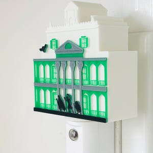 May include: A white and green night light shaped like a mansion. The mansion has green accents, arched windows, and a small rooftop structure. Black silhouettes of figures are in front of the mansion. The night light is plugged into a white wall outlet.