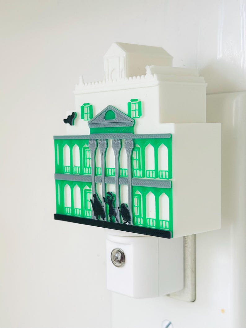 May include: A white and green night light shaped like a mansion. The mansion has green accents, arched windows, and a small rooftop structure. Black silhouettes of figures are in front of the mansion. The night light is plugged into a white wall outlet.