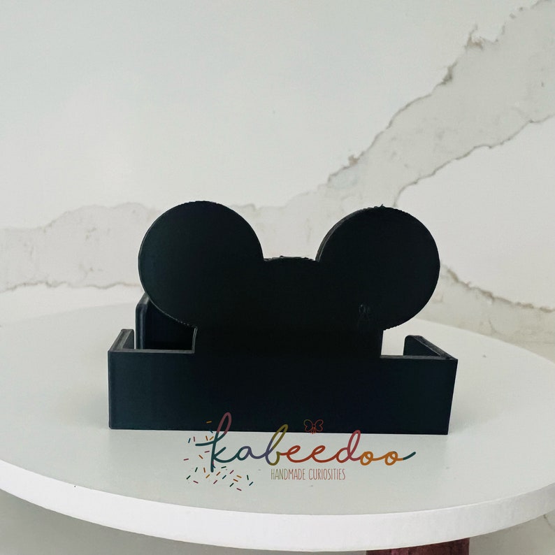 Puede incluir: Juego de posavasos negros con forma de cabeza de personaje de dibujos animados. El juego incluye un soporte y posavasos. El soporte tiene una base rectangular. El texto "kabeedoo handmade curiosities" es visible.