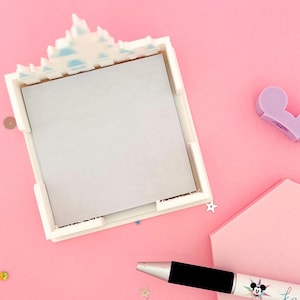 Puede incluir: Un portamemos cuadrado blanco con una parte superior decorativa, que contiene un bloc de notas gris. Un bol&iacute;grafo negro y plateado con un dise&ntilde;o de Mickey Mouse y la palabra "ka" est&aacute; al lado del portamemos. El fondo es rosa con confeti esparcido.
