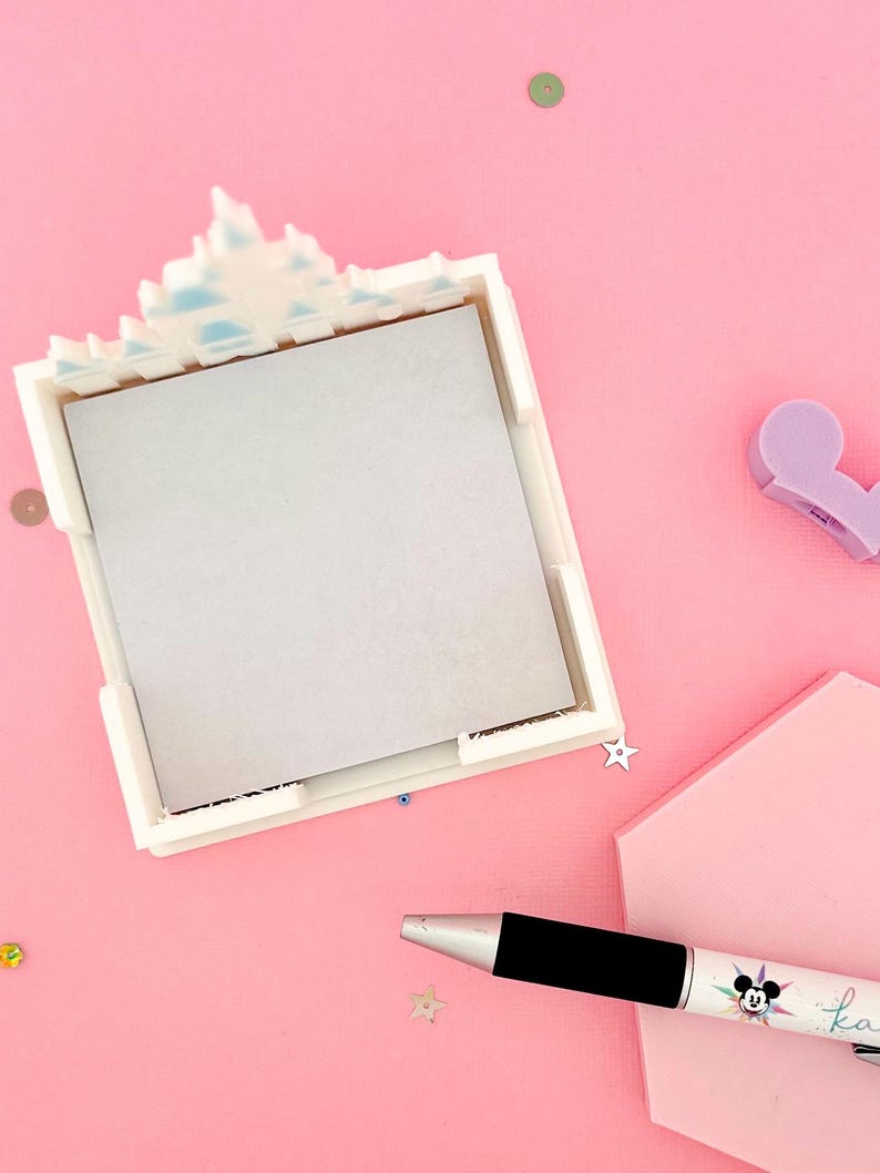 Puede incluir: Un portamemos cuadrado blanco con una parte superior decorativa, que contiene un bloc de notas gris. Un bol&iacute;grafo negro y plateado con un dise&ntilde;o de Mickey Mouse y la palabra "ka" est&aacute; al lado del portamemos. El fondo es rosa con confeti esparcido.