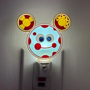 May include: A novelty night light shaped like a cartoon character's face, featuring a light blue face with red polka dots, blue eyes, and three circular shapes. The night light is plugged into a white wall outlet.